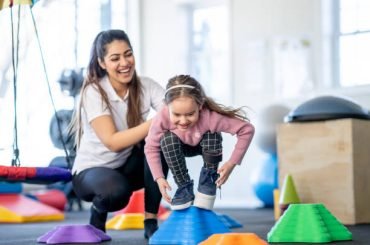 A female physical therapist helps a little girl with a limb difference through a therapy course.  The little girl is balancing on a block as she navigates the colorful course and works on balance with her brace on.